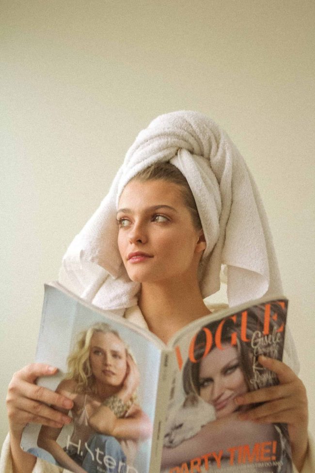 A woman reading a magazine with a bath towel wrapped around her head.